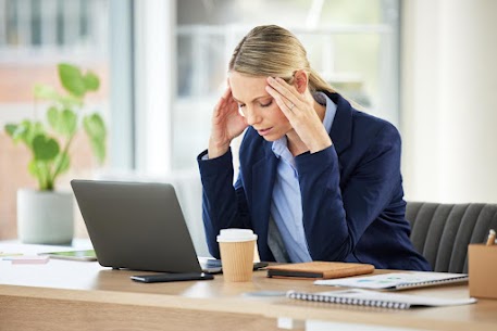 Young woman with blonde hair in a blue suit sitting at a desk in an office holding the sides of her head with tinnitus